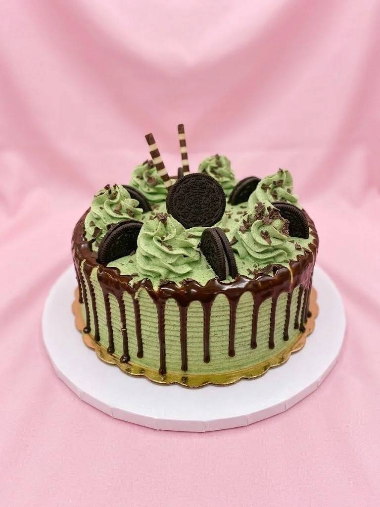 A round cake with green frosting and chocolate drip icing. Decorated with Oreo cookies, green swirls of frosting, striped wafer sticks, and Oreo cookies at the center. Placed on a white cake board against a pink background.