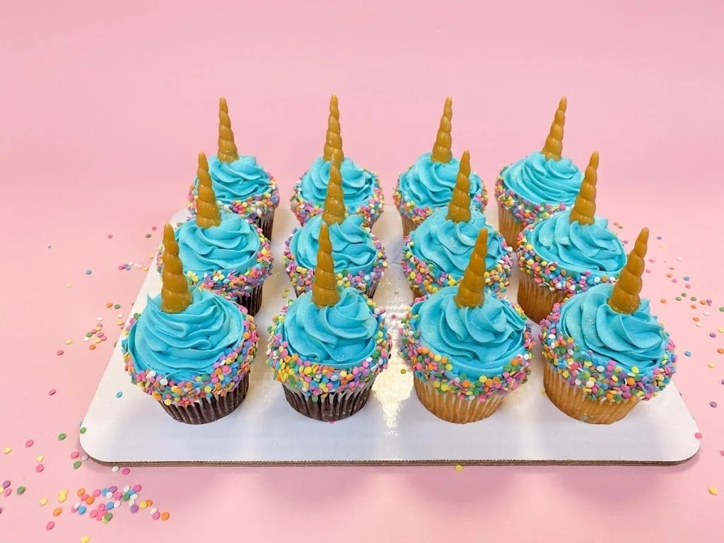 Eleven cupcakes with blue frosting, colorful sprinkles, and gold unicorn horn toppers on a white rectangular tray, against a pink background.