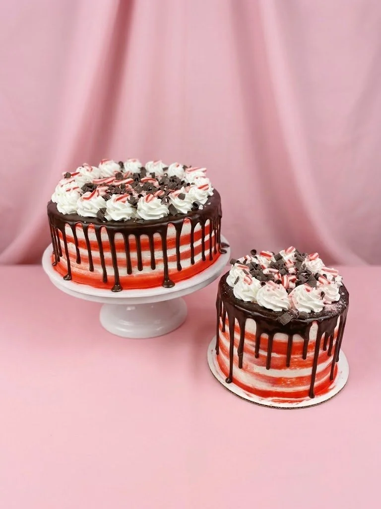 Two decorated cakes with pink and white layers, topped with whipped cream, chocolate shavings, and pink and white striped candies, placed on a pink surface with a pink background.