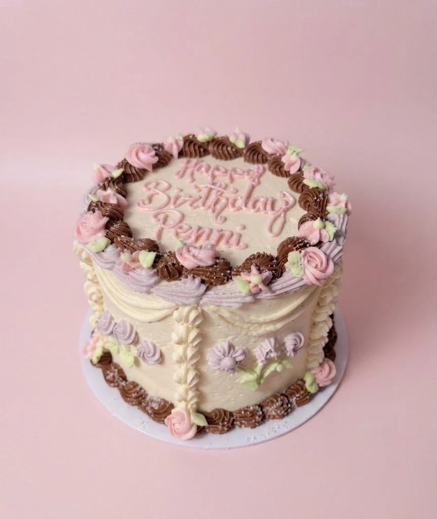 Cake with pink and brown frosting decorations, pink and green flower accents, and the message "Happy Birthday Penni" written on top.