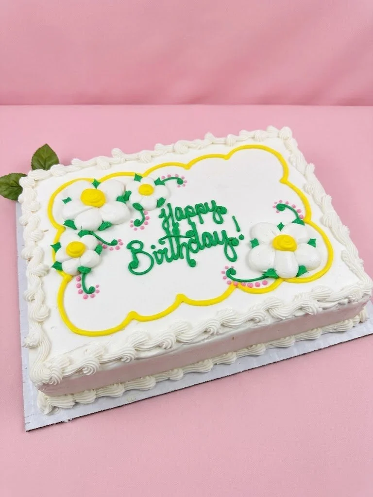 Rectangular birthday cake with white frosting, decorated with white flowers with yellow centers, green leaves, and pink dots, on a pink background, with green "Happy Birthday!" message.