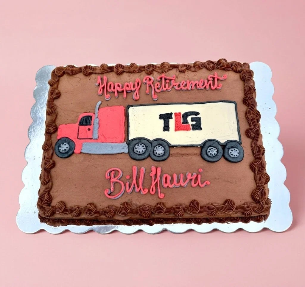 A decorated birthday cake with chocolate frosting, pink icing writing that says 'Happy Retirement Bill Hauri', and a truck design with 'TLG' on the trailer, on a pink background.