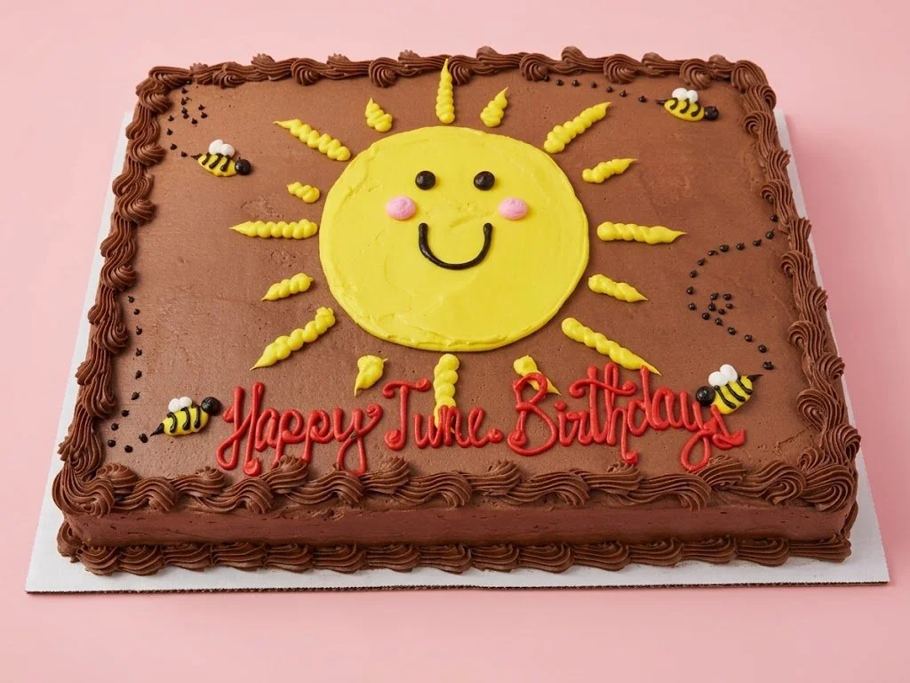 Chocolate birthday cake with a smiling sun face made of yellow frosting on top, surrounded by bees and the words "Happy Tune Birthday" written in red icing. The cake has a decorative border of chocolate frosting.