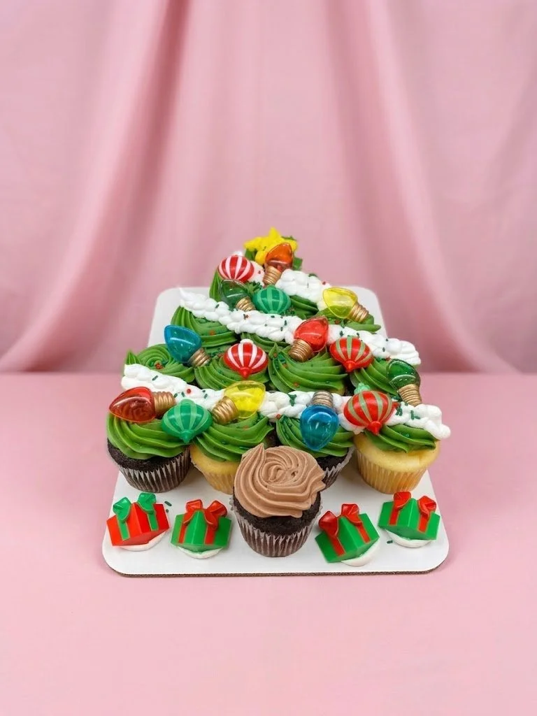 Christmas-themed cupcakes and cake with holiday decorations on a white tray against a pink background.