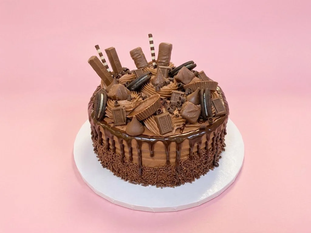 Chocolate cake decorated with various chocolate candies, biscuits, and chocolate sticks on top, with chocolate drizzle on the sides, on a white cake board against a pink background.