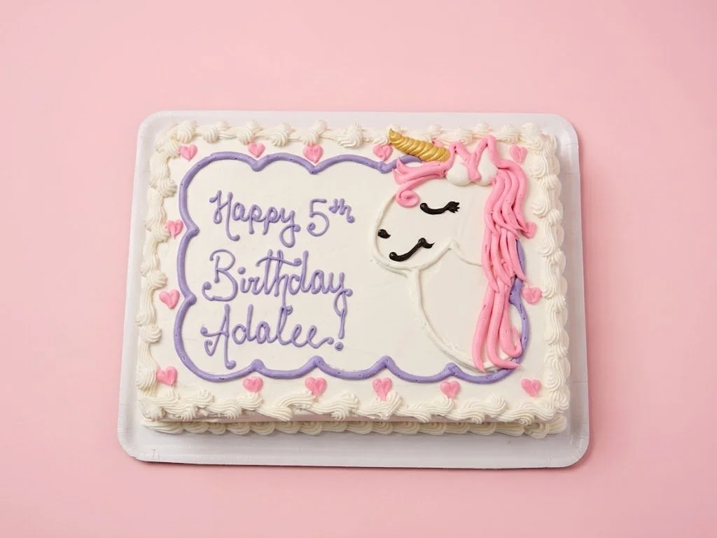 A rectangular birthday cake decorated with a unicorn face, pink and purple icing, small pink hearts, and a message that reads "Happy 5th Birthday Adalee!" on a pink background.