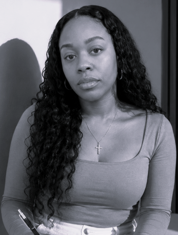 Black and white photo of a young woman with long curly hair wearing a top and a cross necklace, sitting indoors