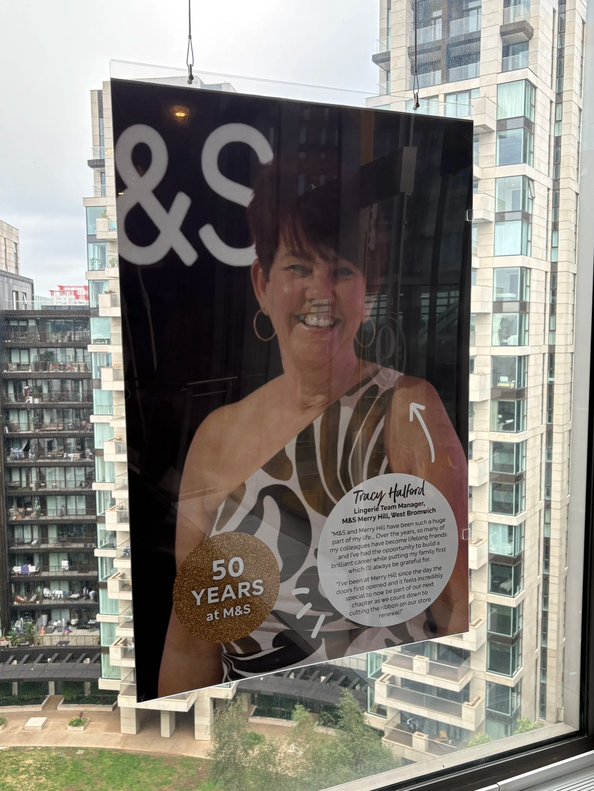 A window with a poster celebrating 50 years at M&S. The poster features a smiling woman with short dark hair wearing hoop earrings, and contains text about her career and achievements, including her role as a lingerie team manager.