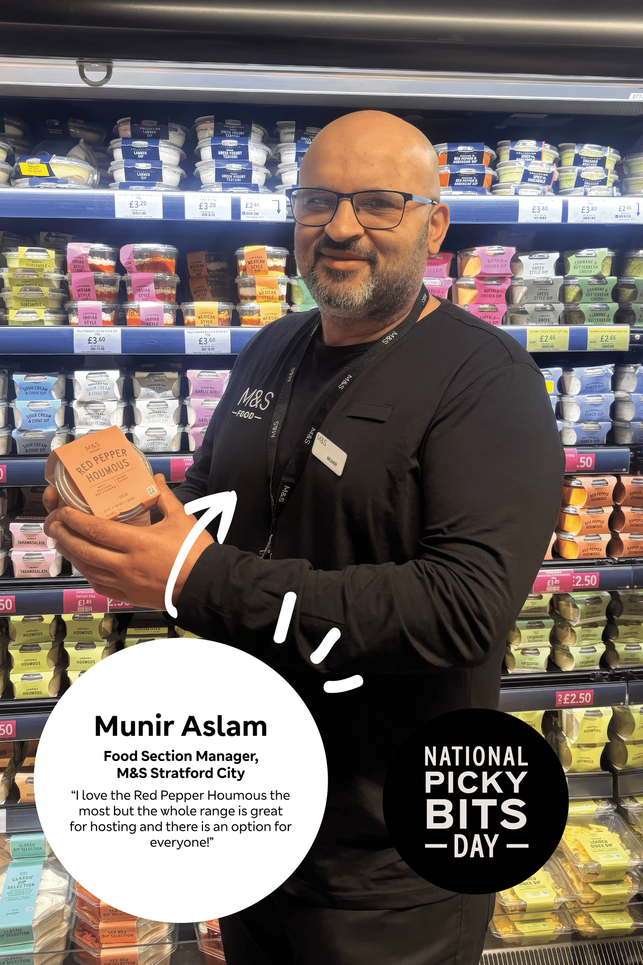 A man with glasses and a beard holding a container of Red Pepper Houmous in a grocery store aisle with shelves of various dips behind him.