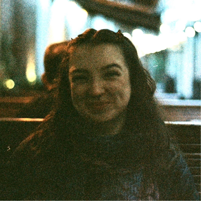 A woman with dark, wavy hair smiling in a dimly lit outdoor setting.