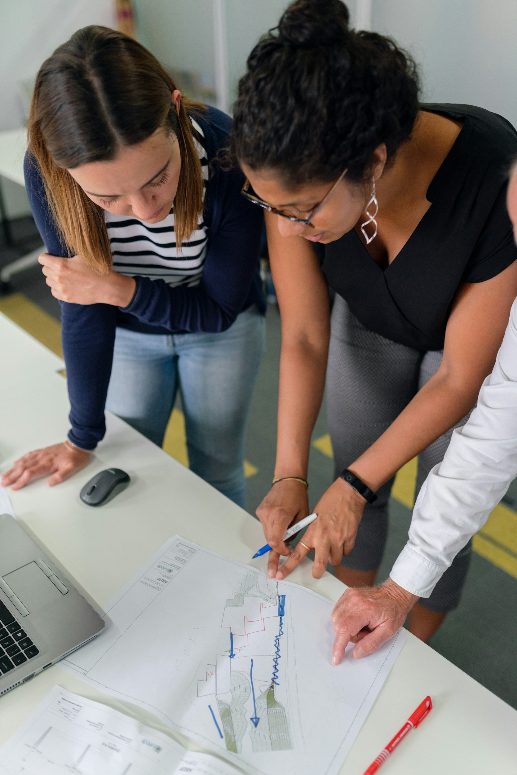 Two women are working together over architectural plans and maps on a table. They are pointing at specific areas on the plans, discussing them, with a laptop and writing utensils nearby.