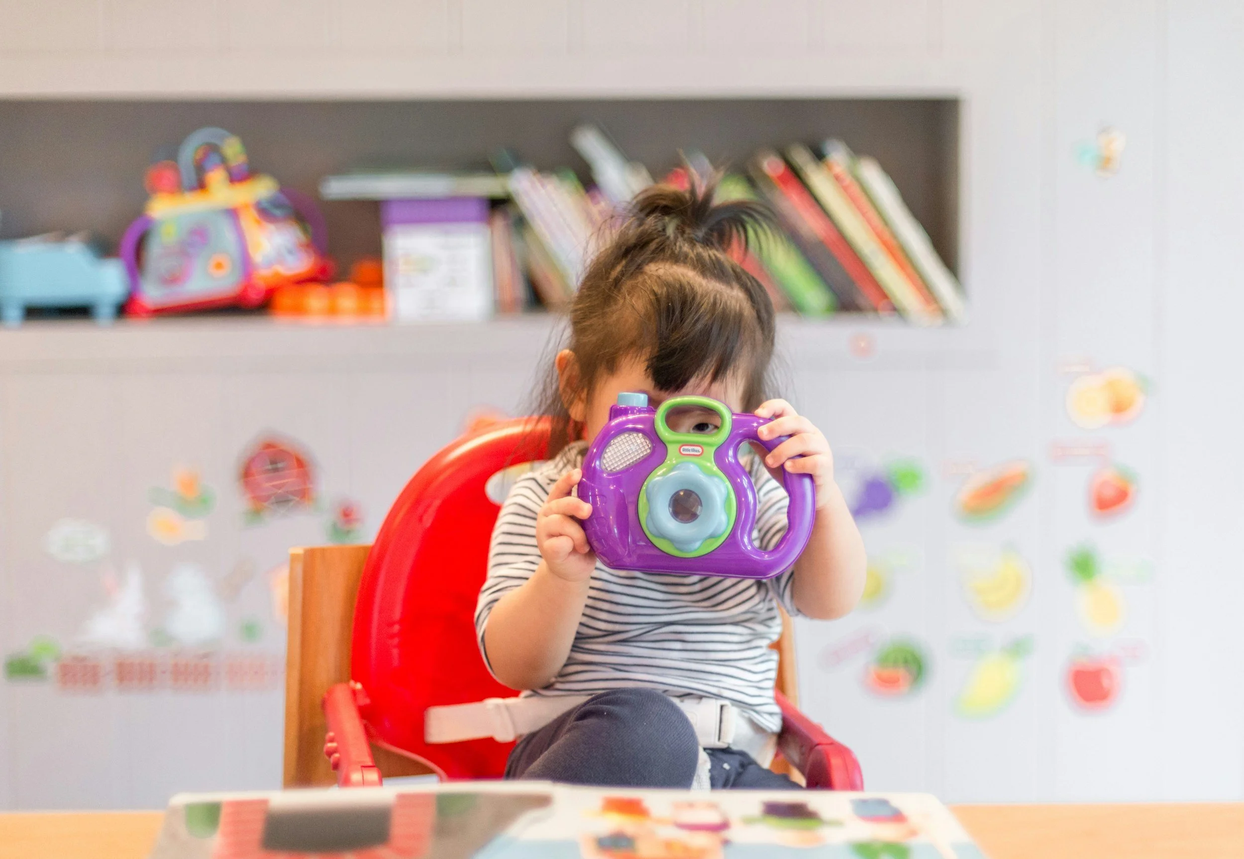 A young girl with dark hair in a messy ponytail is sitting in a red chair at a table, holding a purple and green toy camera up to her face, partially obscuring it. In the background, there's a grey storage unit with colorful books and toys on the shelf, and some colorful stickers on the wall.