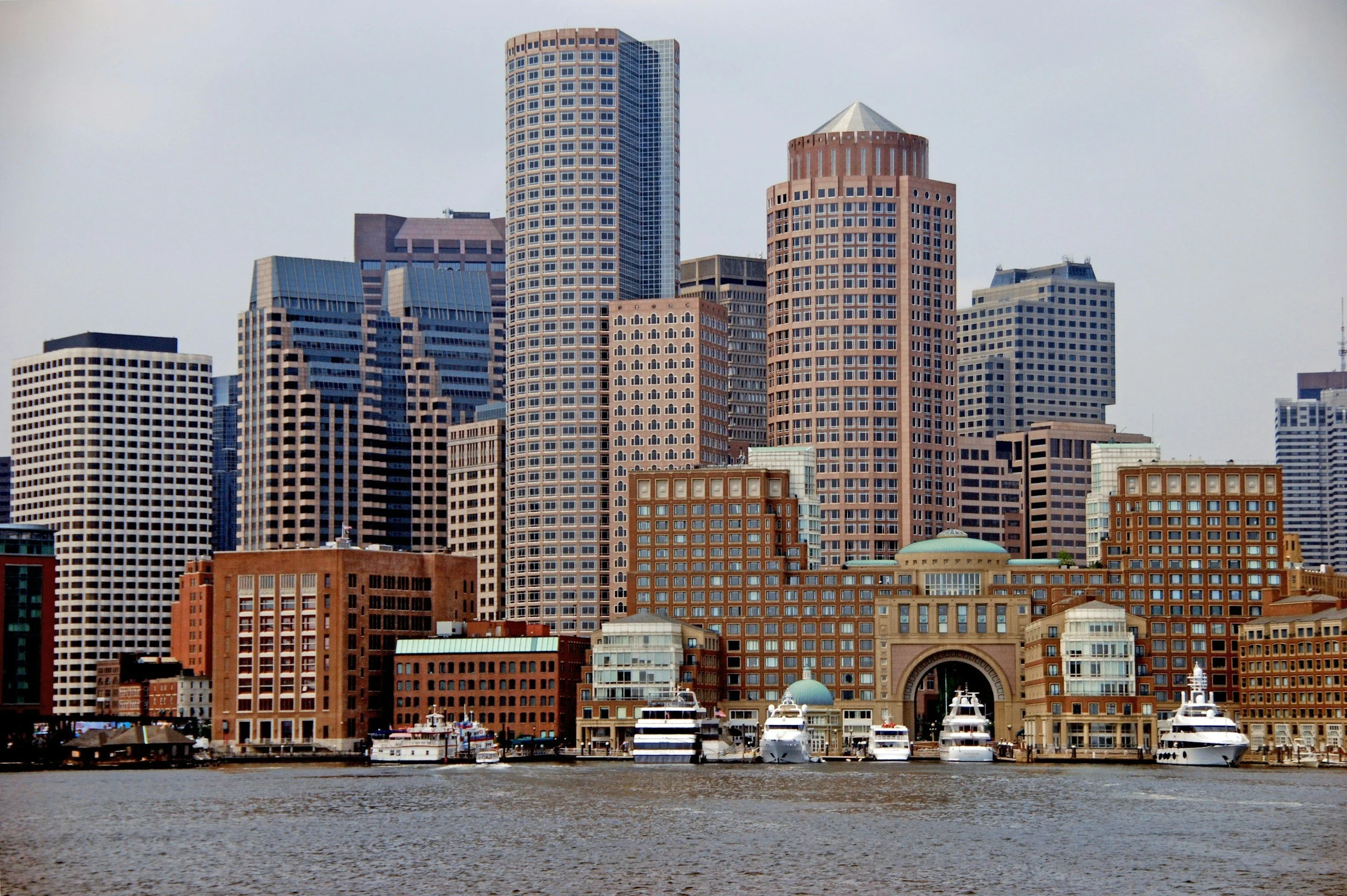A city skyline with multiple tall buildings and yachts docked in the water in the foreground.