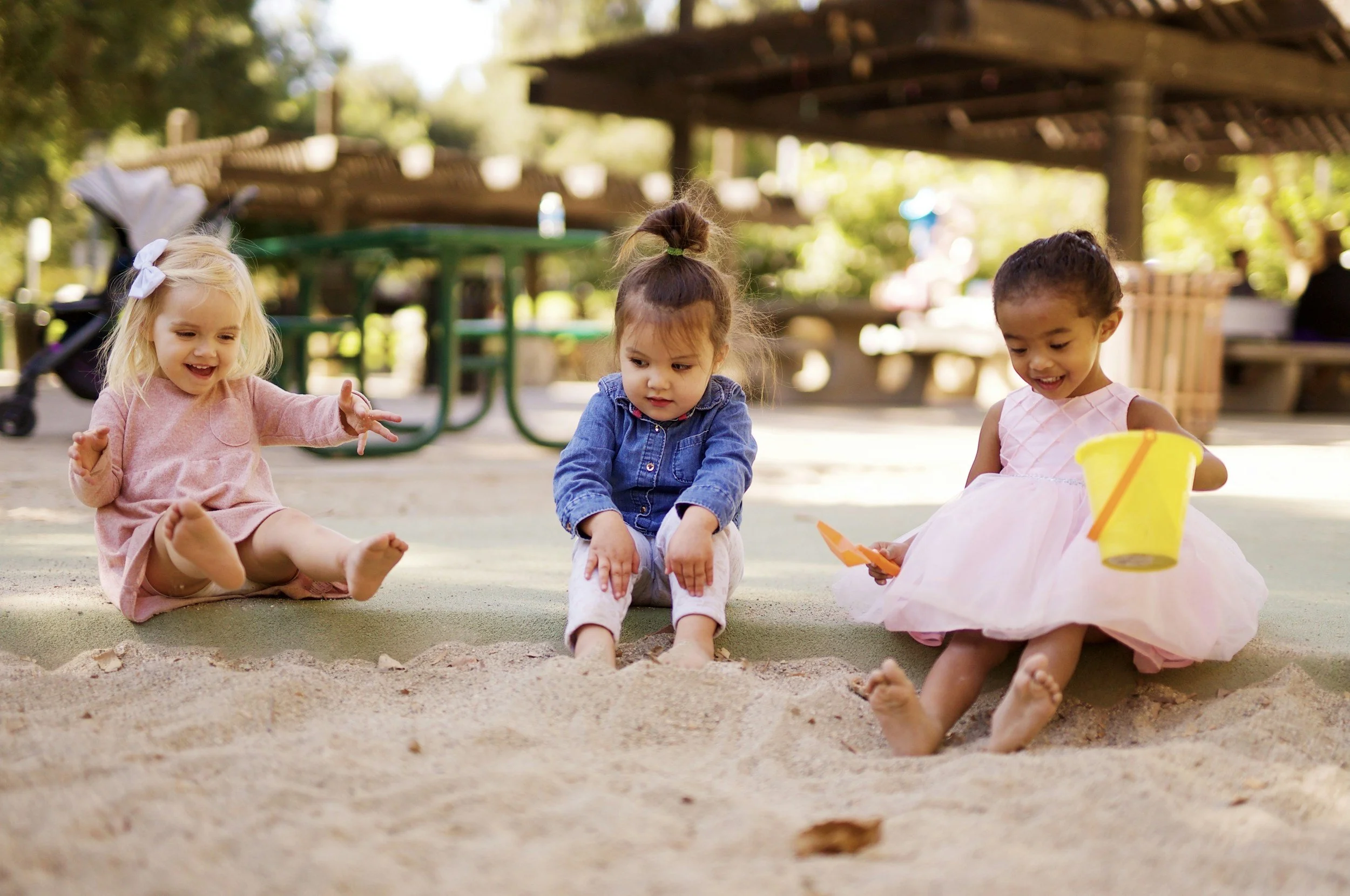 Three young girls playing in a sandbox at a park, with trees and picnic tables in the background.