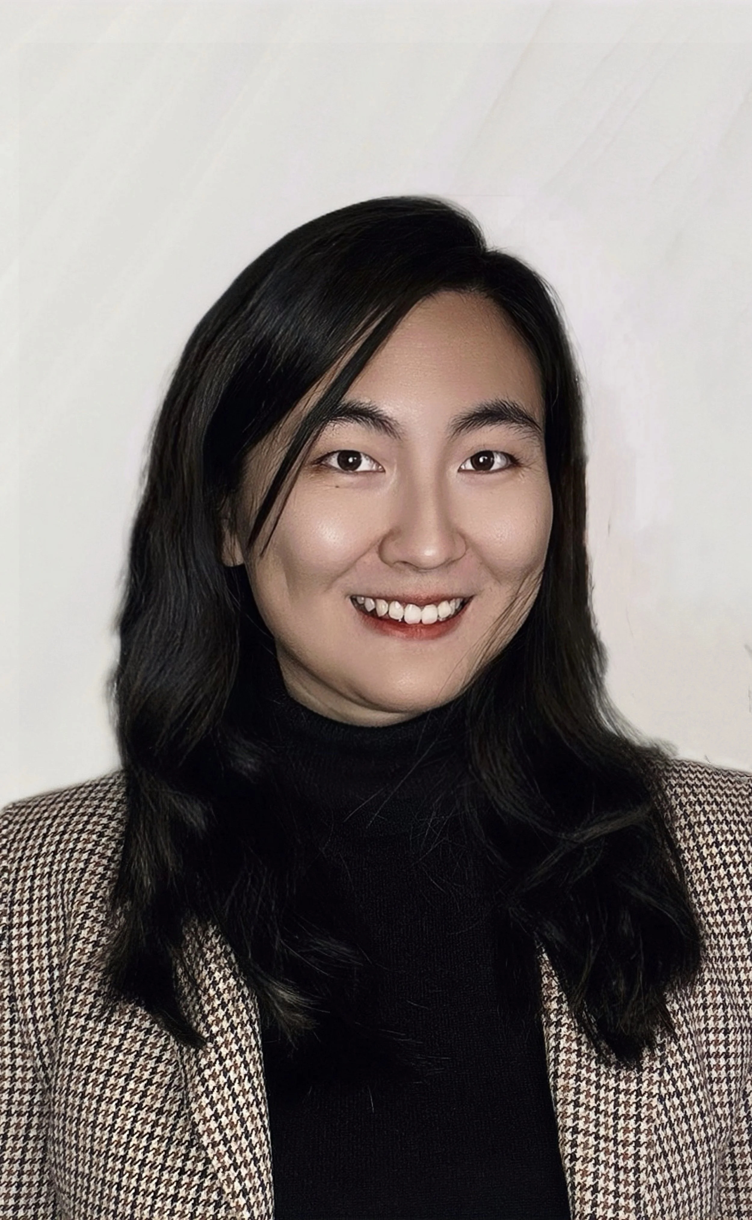 Portrait of a woman with long black hair, wearing a black turtleneck and a brown plaid blazer, smiling against a plain white background.