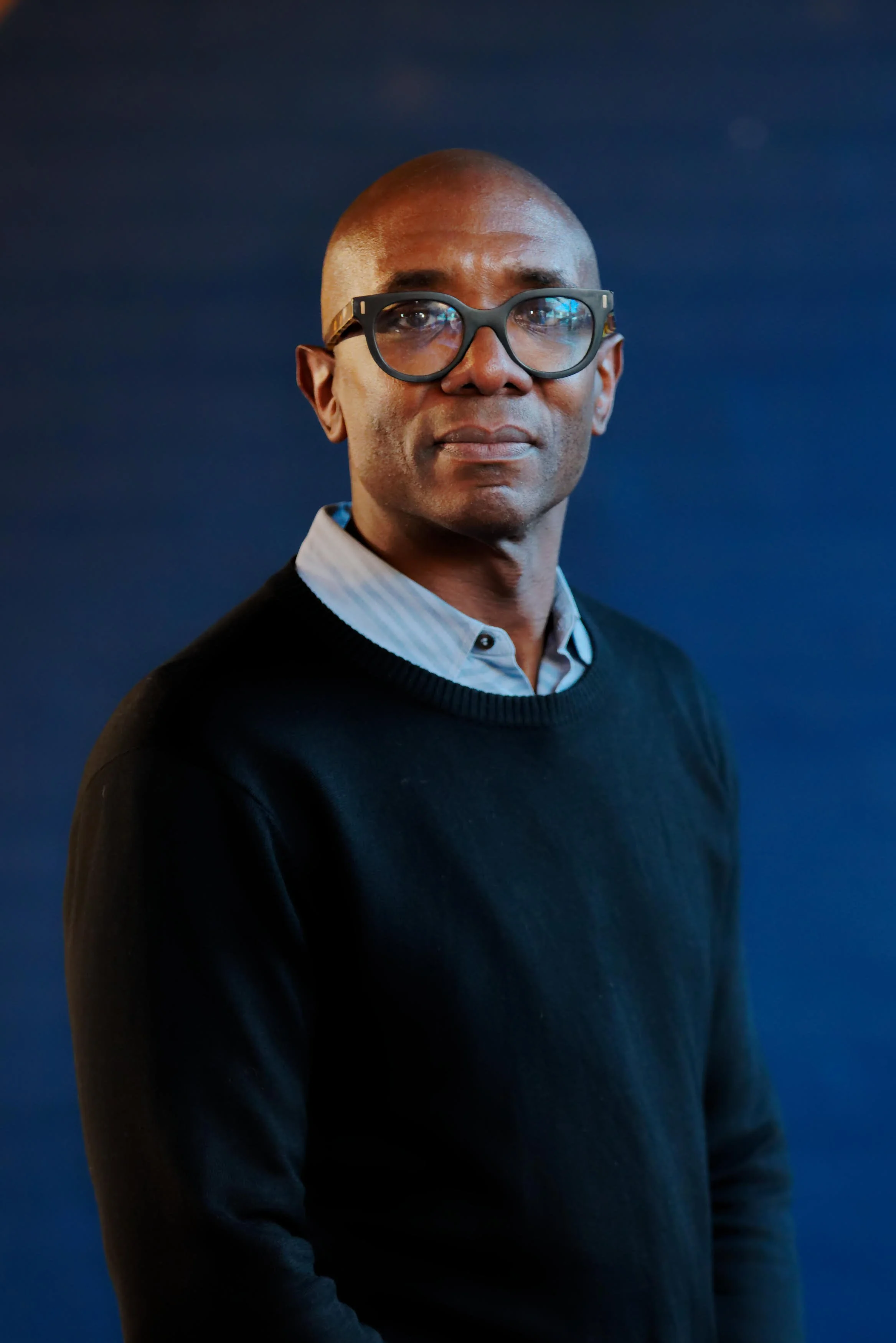 Portrait Bert Stemarthe wearing glasses, light blue collared shirt and navy blue jumper, standing against a dark blue background.