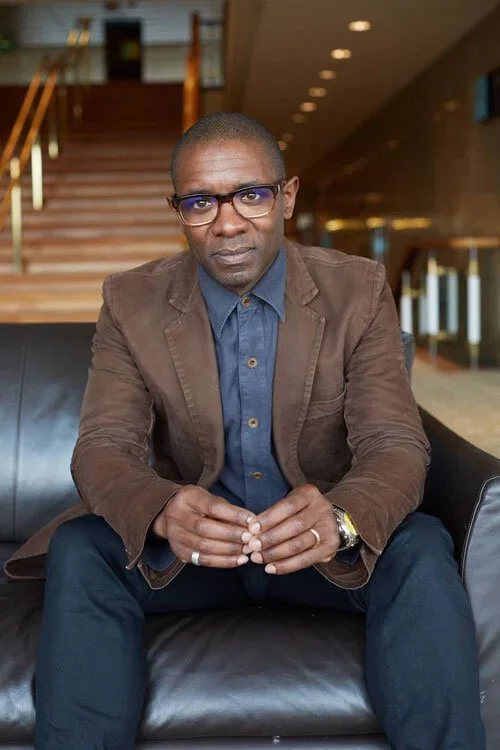 Bert Stemarthe (therapist, executive coach and business psychologist) sitting on a black leather couch inside a modern building, wearing glasses, a brown blazer, blue shirt and dark trousers, with hands clasped, looking directly at camera.