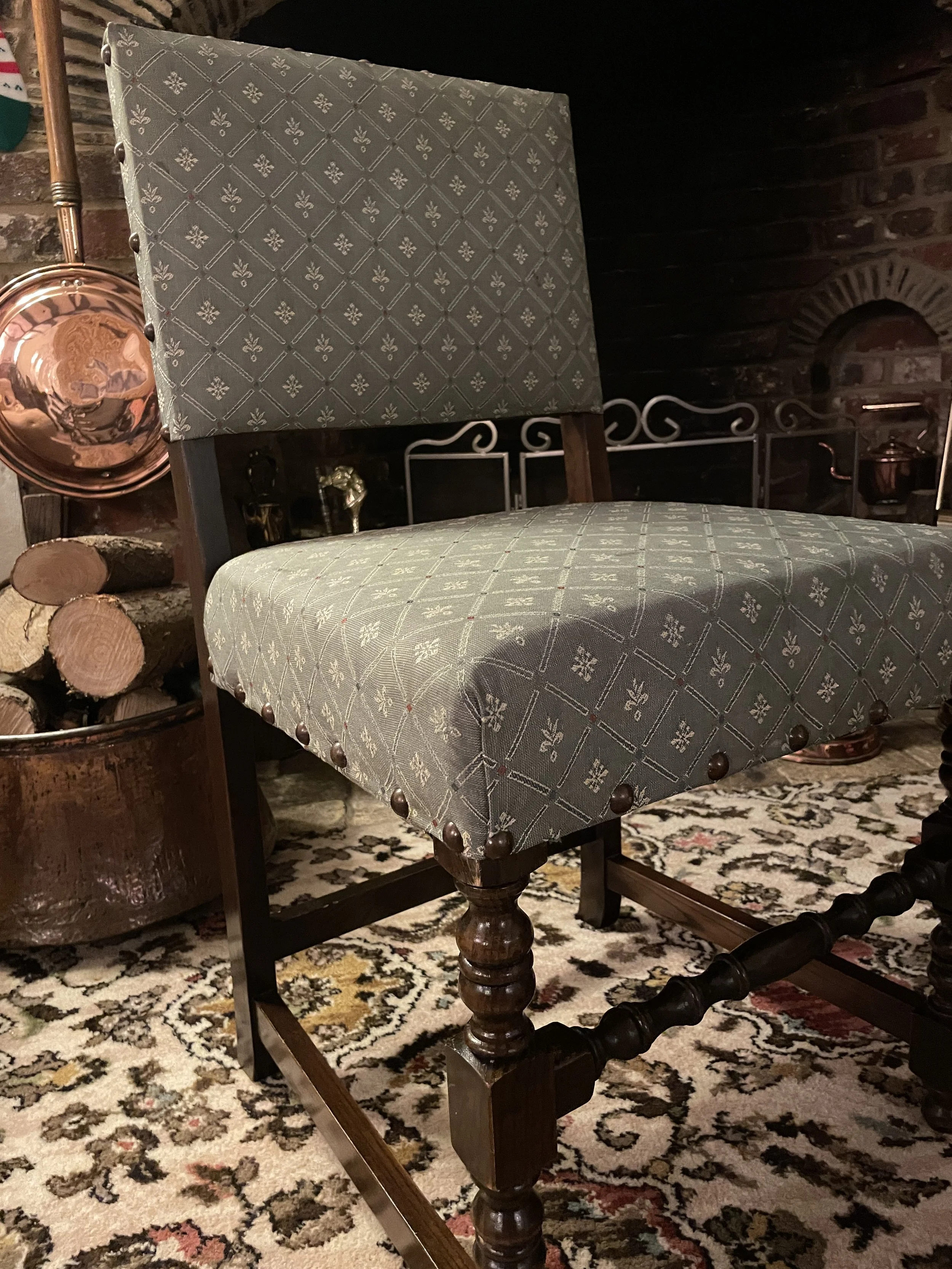 A green dining chair stands infron tof a wide brick hearth, a basket of logs and highly polished brass work hang in the background.