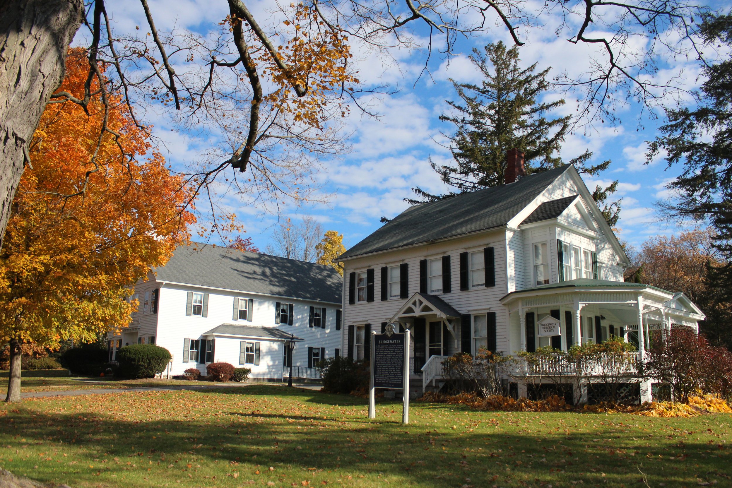 The Elijah Peck house, located in Bridgewater, CT. Home of the Bridgewater Historical Society, it has black shutters and a porch, surrounded by trees with fall foliage, under a partly cloudy sky.