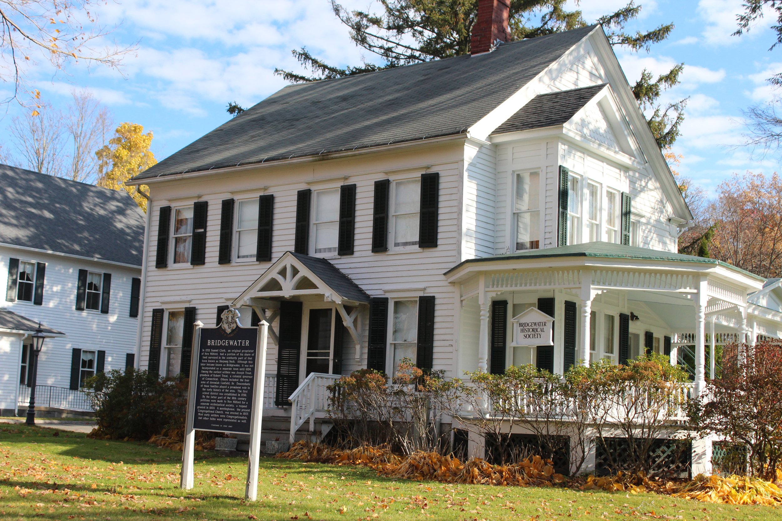 The Elijah Peck House. A white two-story house that is the home of the Bridgewater Historical Society, with black shutters and a porch, surrounded by autumn-colored trees and fallen leaves, with a historical society sign in front.