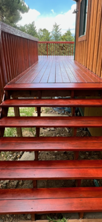 Red wooden porch with steps leading up to a small deck, surrounded by green trees and a cloudy sky.