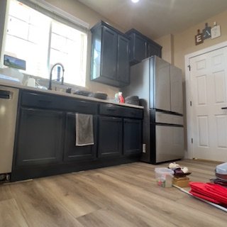 A kitchen with black cabinets, a stainless steel refrigerator, a window above the sink, and various items on the counter and floor.
