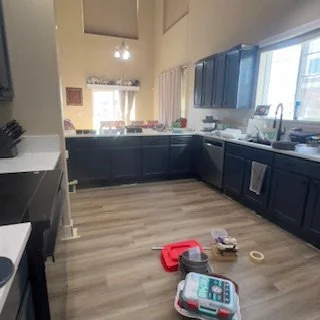 Empty kitchen with dark blue cabinets, light wood flooring, and a window over the sink. Countertop clutter includes a red cutting board, utensils, and small items.