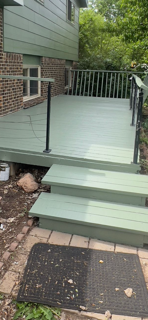 Outdoor deck with steps leading up to a house with green siding and brick foundation, surrounded by trees.