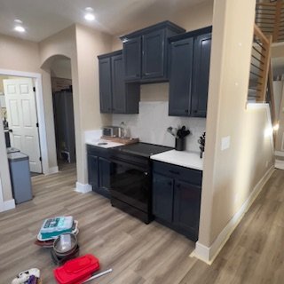 Kitchen with navy blue cabinets, white countertops, and wooden flooring. Items on the floor include a stack of papers, a pet food bowl, a red container, and a gray container. Part of a staircase is visible on the right.