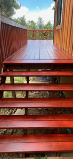 Red wooden deck with steps leading up to a fenced porch, surrounded by green trees.