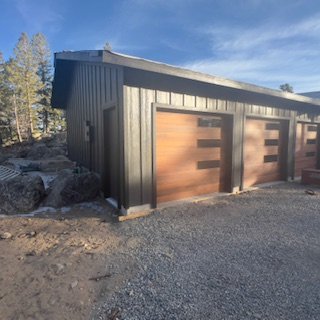 A black metal building with three garage doors featuring a horizontal cutout design, set against a background of trees and a blue sky.