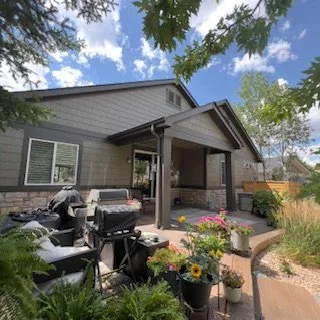 Backyard patio area with outdoor furniture, barbecue grill, potted flowers, and a house with gray siding under a partly cloudy sky.