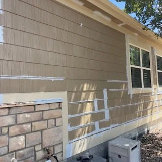 Side view of a house exterior with beige siding, a window, and a section of brickwork near the bottom, with shadows cast on the wall and some construction tape on the siding.