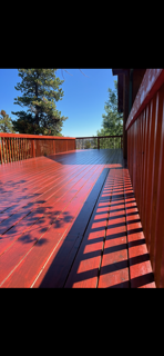 Wooden deck with red railing and shadow patterns, overlooking trees under a clear blue sky.