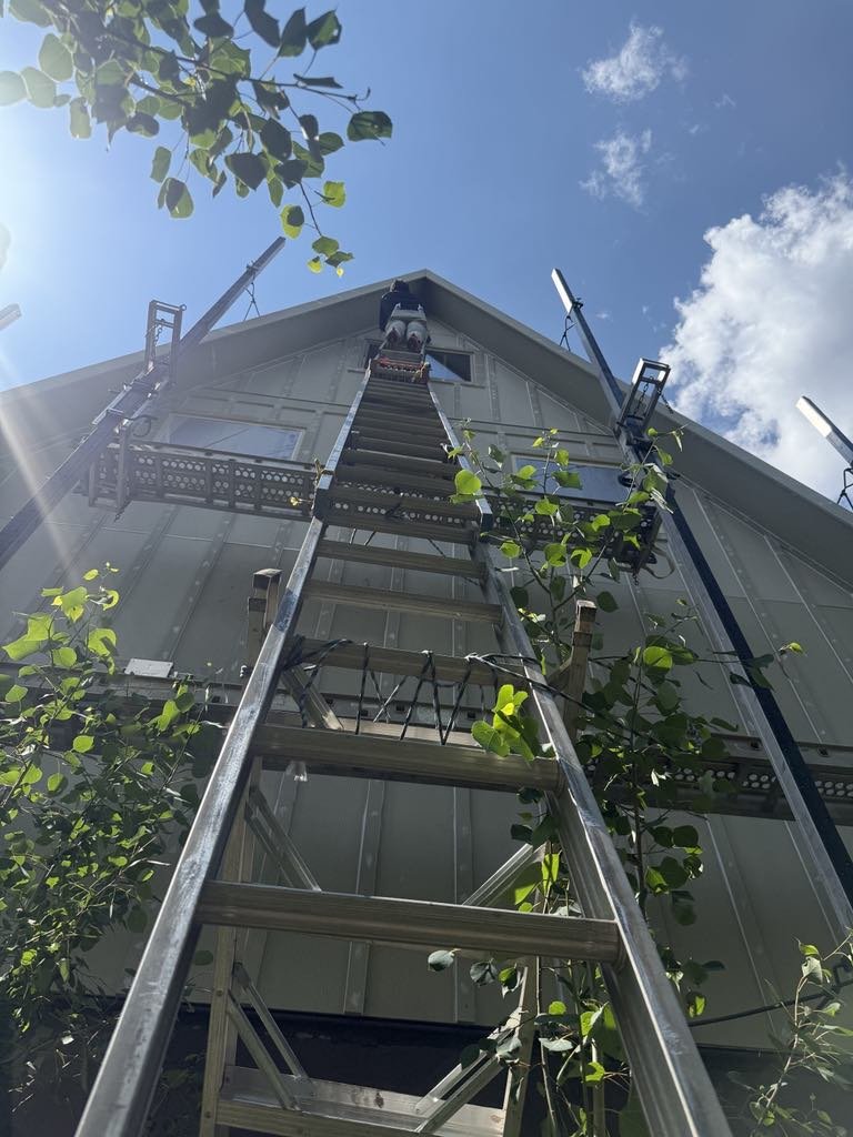 Applying siding on a house under a blue sky with some clouds, with a tall ladder and equipment nearby.