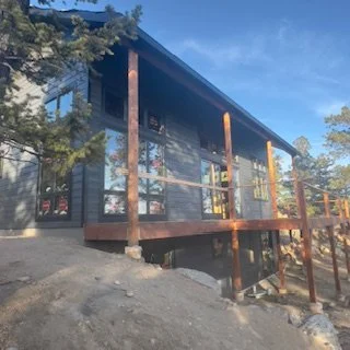 A modern house with large glass windows and a wooden deck, built on a rocky terrain surrounded by trees, with a clear blue sky above.