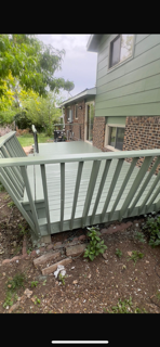 A white wooden railing in the foreground with a garden and sidewalk in the background, neighboring houses, trees, and a person walking along the sidewalk.