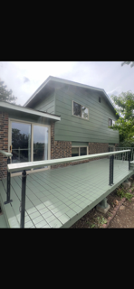 View of a modern two-story house with green siding and brick accents, featuring a balcony with a metal railing and large windows.