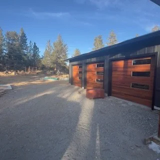 A modern garage or shed with wooden doors located on a gravel driveway, surrounded by trees under a blue sky with some clouds.