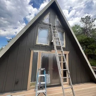 A black A-frame house with a ladder leaning against the front, and a small blue step stool in front. The house has a door and windows on the front side.