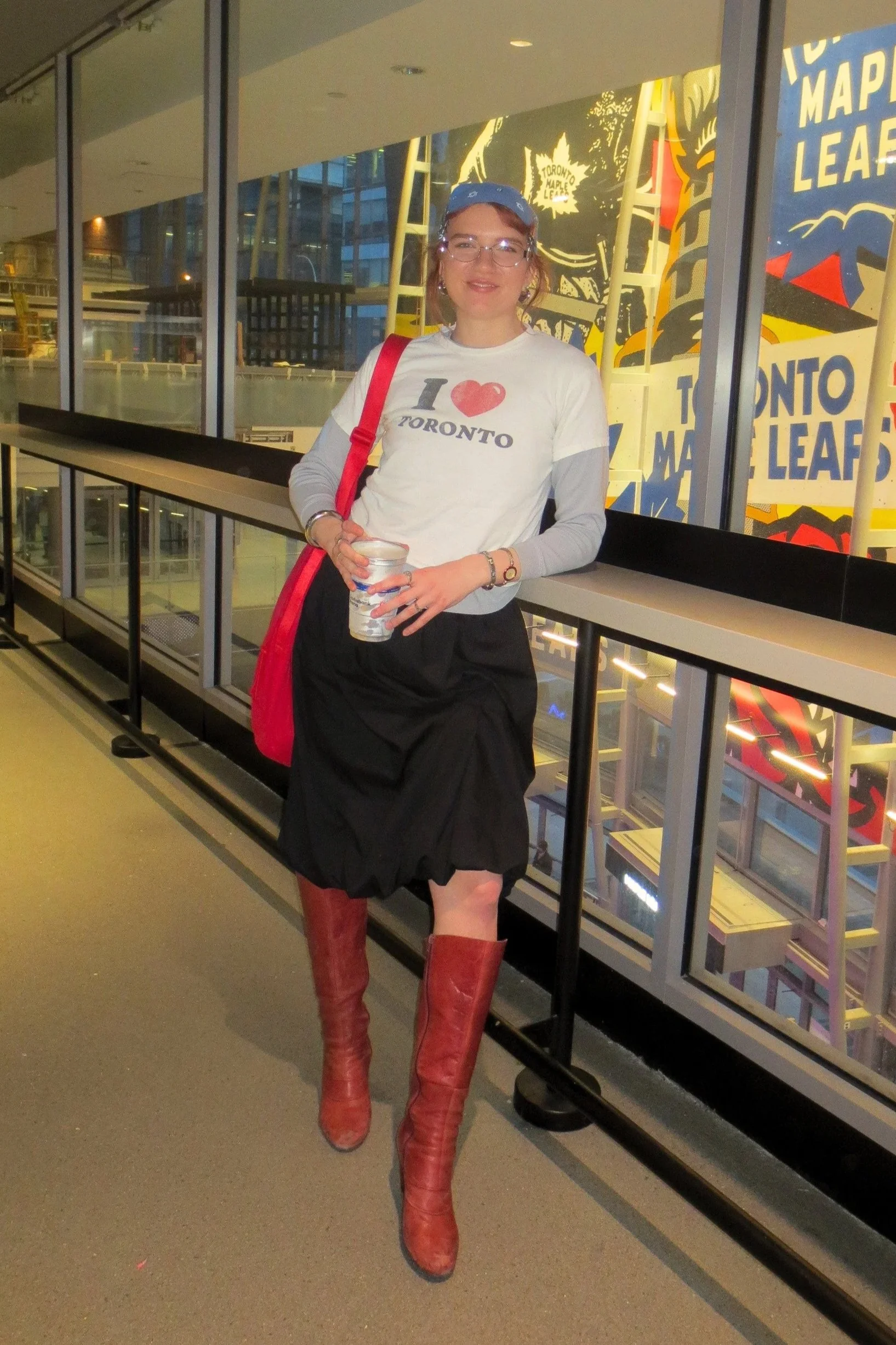 A person standing indoors on a balcony, wearing a white 'I love Toronto' shirt, black skirt, red thigh-high boots, glasses, and a blue bandana, holding a cup.