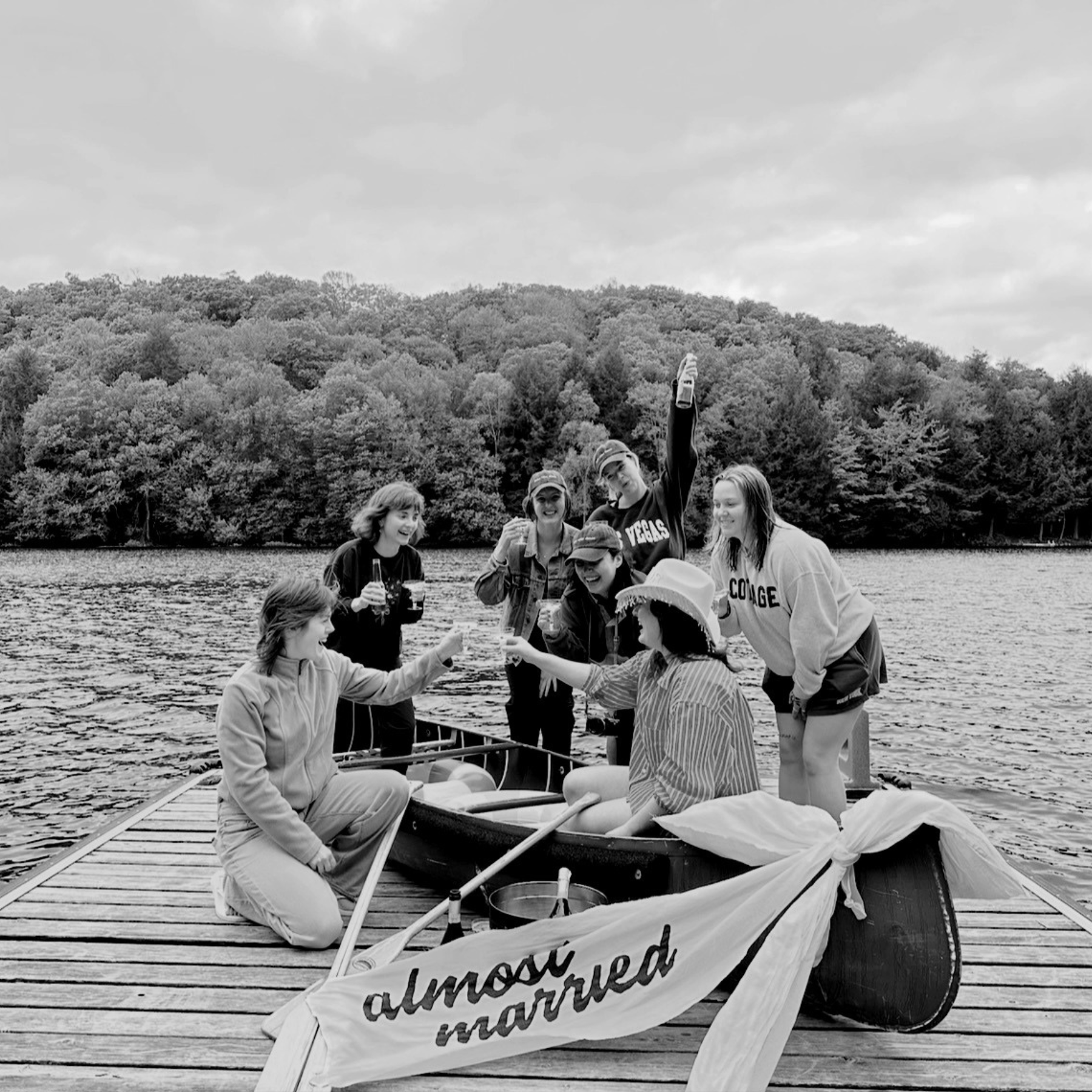 A group of young people on a canoe on a dock with a lake and forest in the background. Some are smiling and holding drinks, others are sitting or standing, wearing casual clothes and hats.
