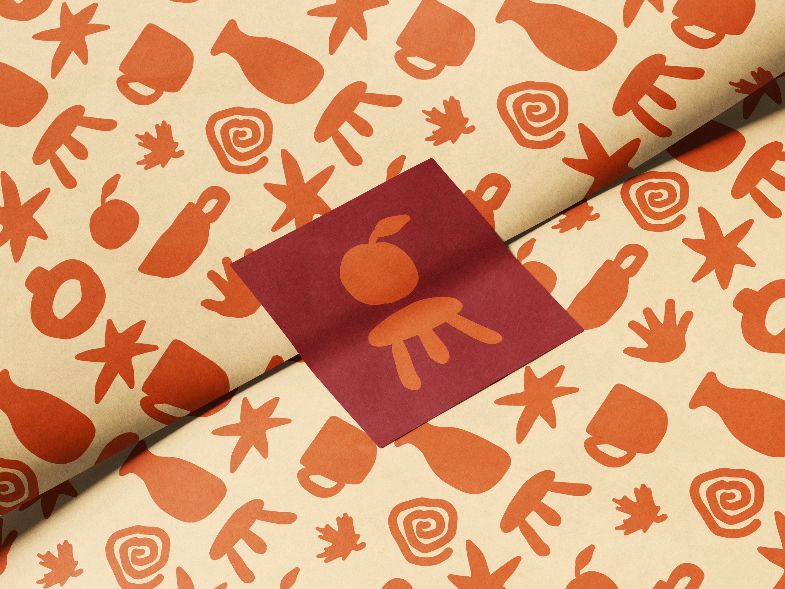 Light yellow tissue paper folded and held together by a dark red branded packaging sticker. The paper and sticker both feature orange hand-drawn silhouette icons of: spirals, vases, teacups, oranges, stars, hands, maple leaves, stools, mugs.
