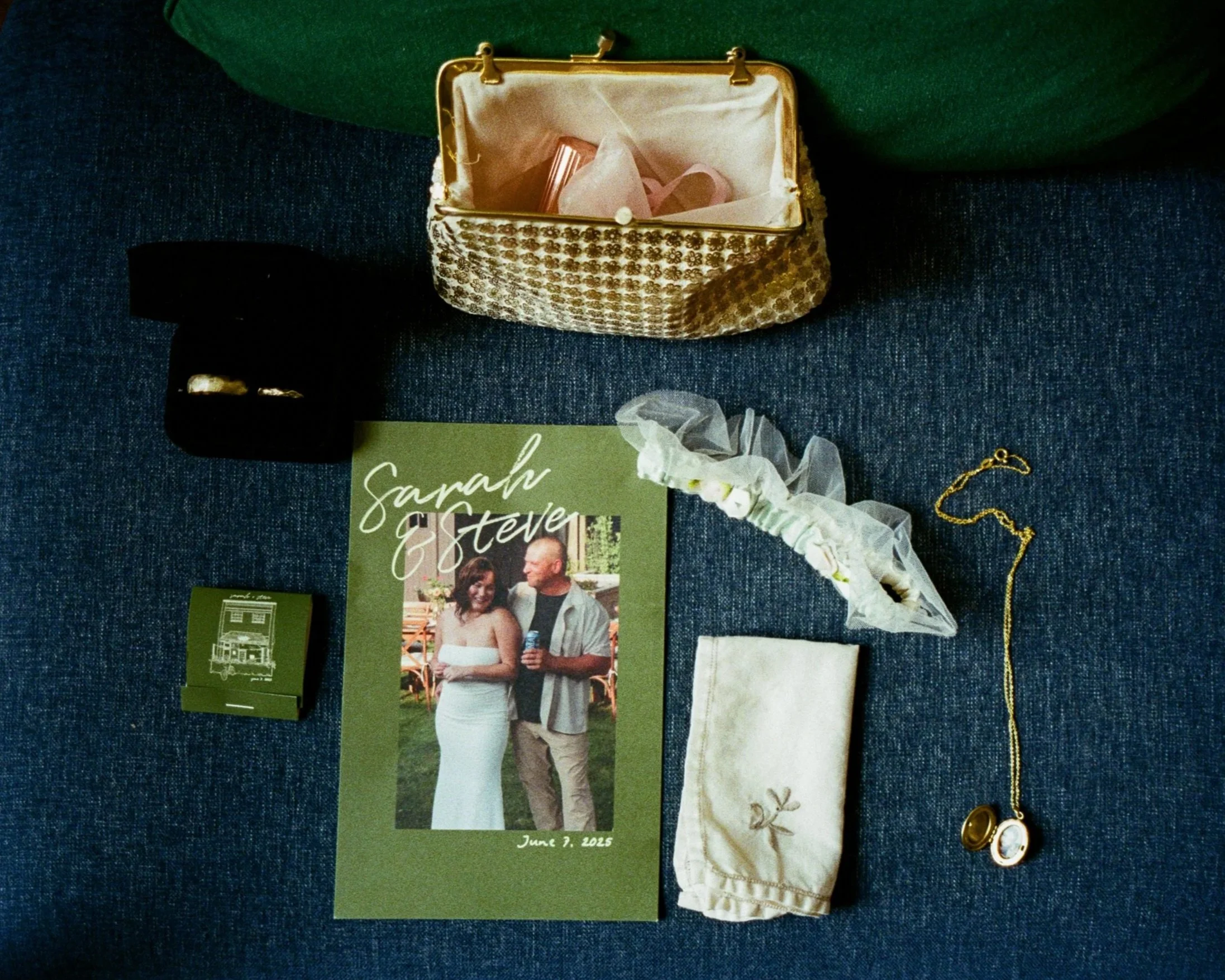 A wedding invitation and a photograph of a smiling couple are displayed on a blue fabric surface. The invitation is green with white cursive text and a photo of a man and woman. The photo on the invitation shows the couple standing outdoors.