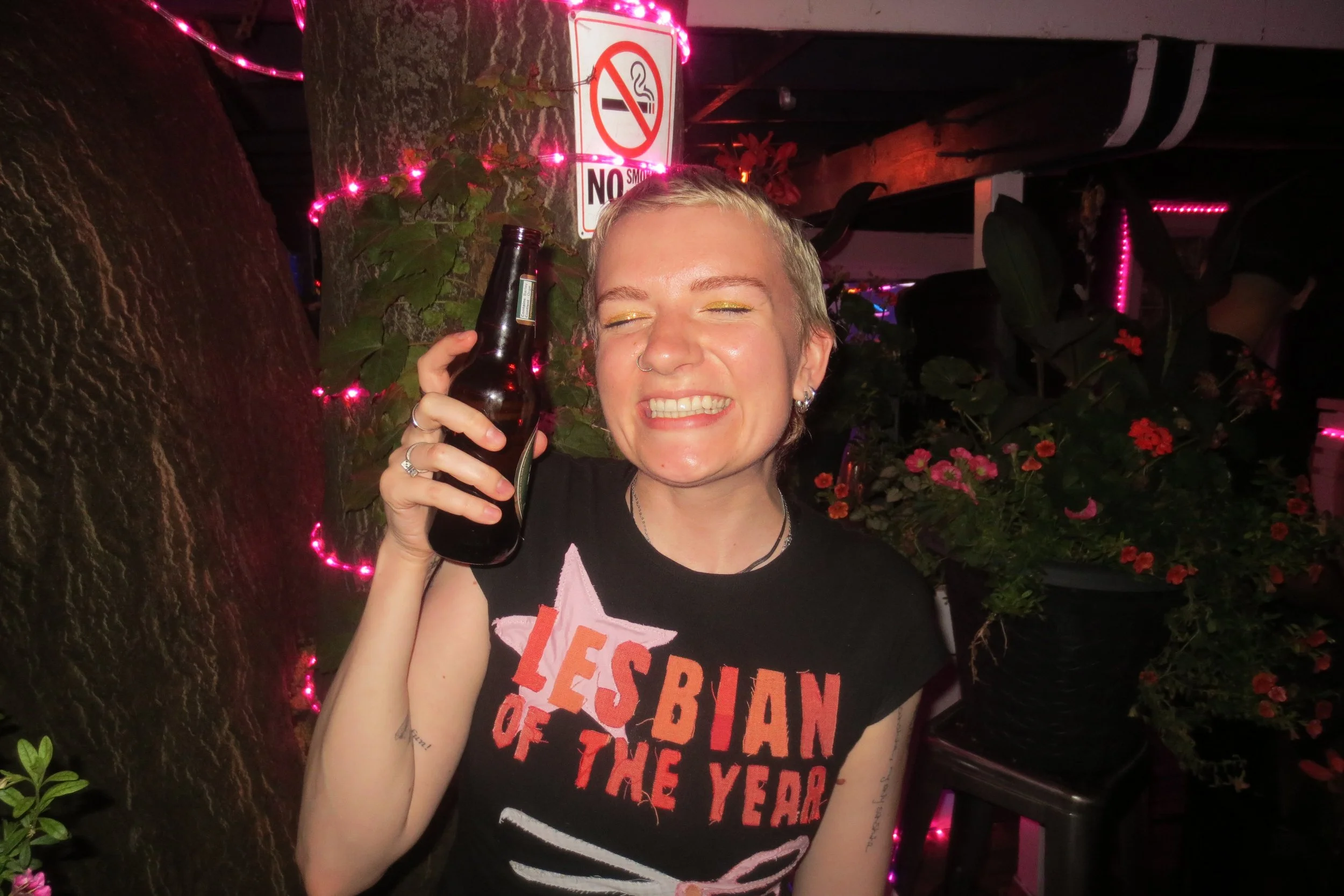 A person with short blonde hair smiling with eyes closed, holding a beer bottle.
