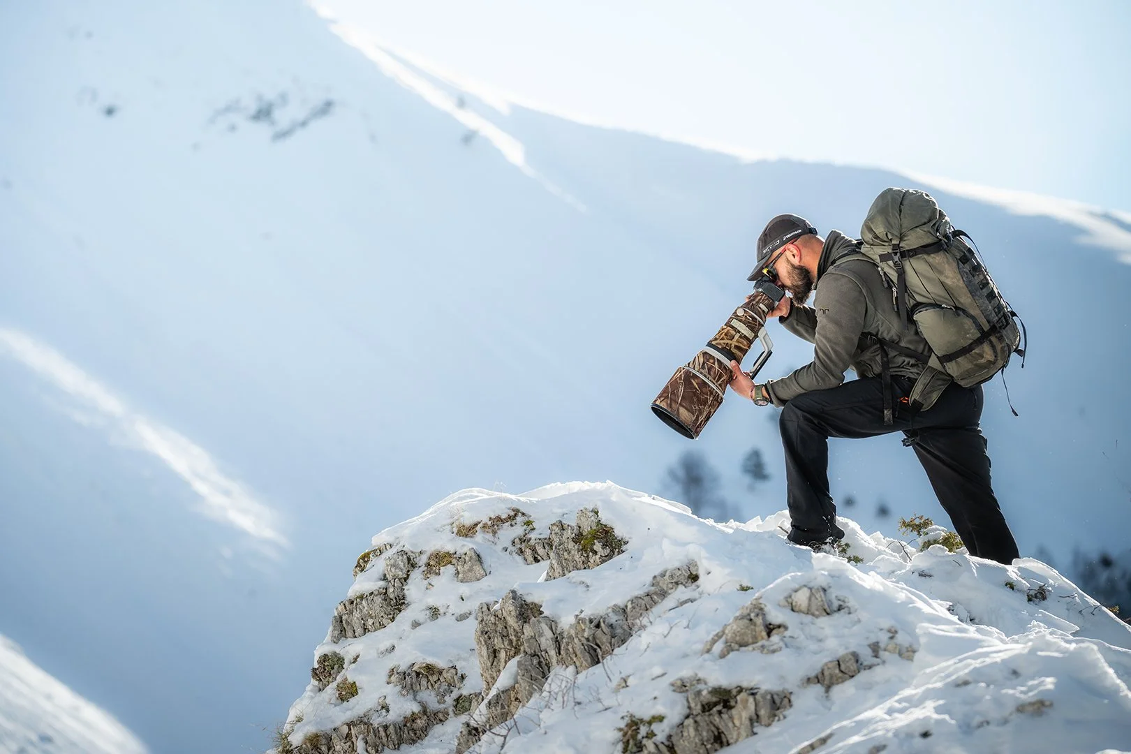 Man with backpack and hiking jacket photographs wildlife on a snowy mountain with telephoto lens