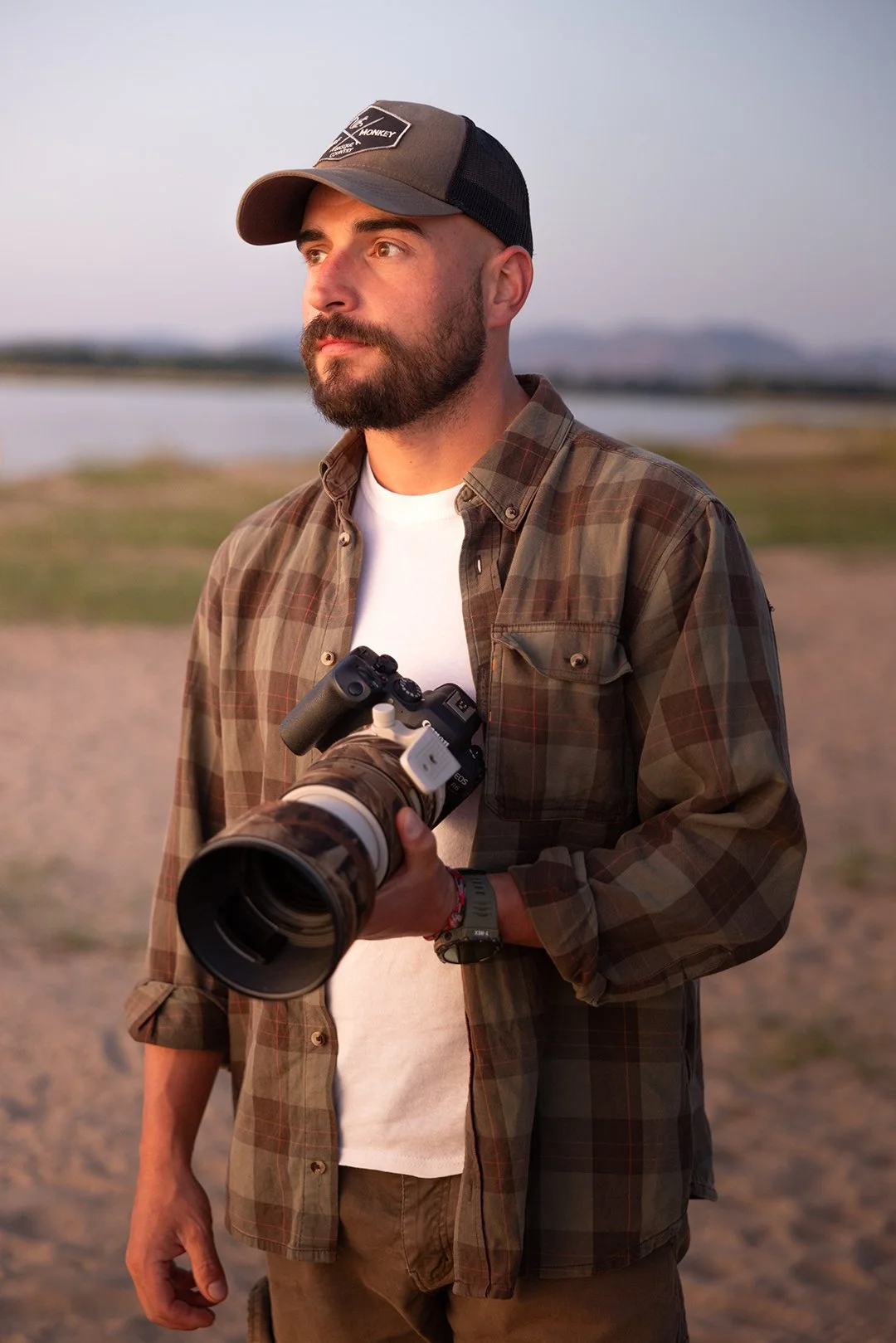 Un uomo con barba indossa una maglietta bianca e una camicia a quadri con cappellino mentre tiene una macchina fotografica con un grande teleobiettivo. Sfondo di un paesaggio naturale al tramonto.