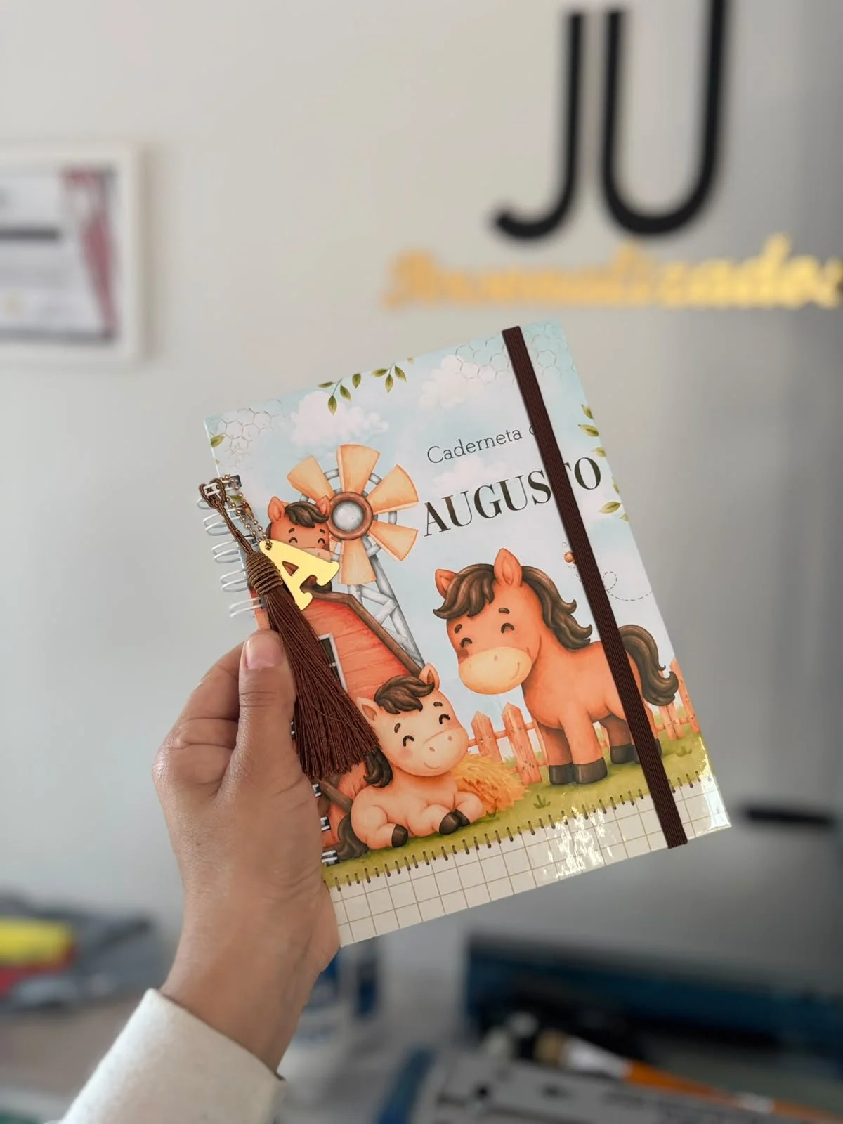 Caderno de rotina para o mês de agosto, com capa decorada com desenhos de cavalos jovens e uma roda de moinho de vento. Um laço marrom com um espiral e uma letra 'A' dourada estão presos ao caderno.