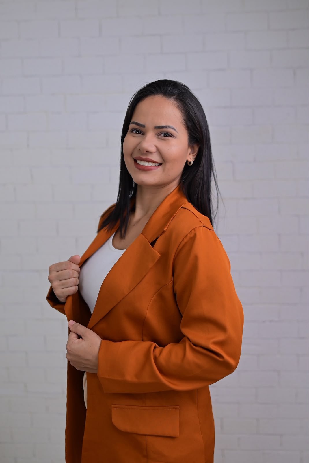 Mulher sorridente com cabelo escuro, vestindo blazer laranja e camiseta branca, segurando a lapela do blazer, posando contra fundo de parede de tijolos brancos