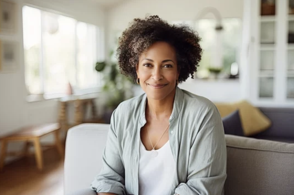 A woman with curly hair smiling and sitting in a cozy living room with natural light.